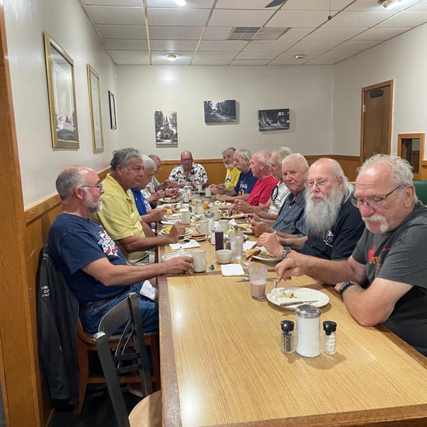 Group of men eating breakfast at a long table in a casual diner setting.